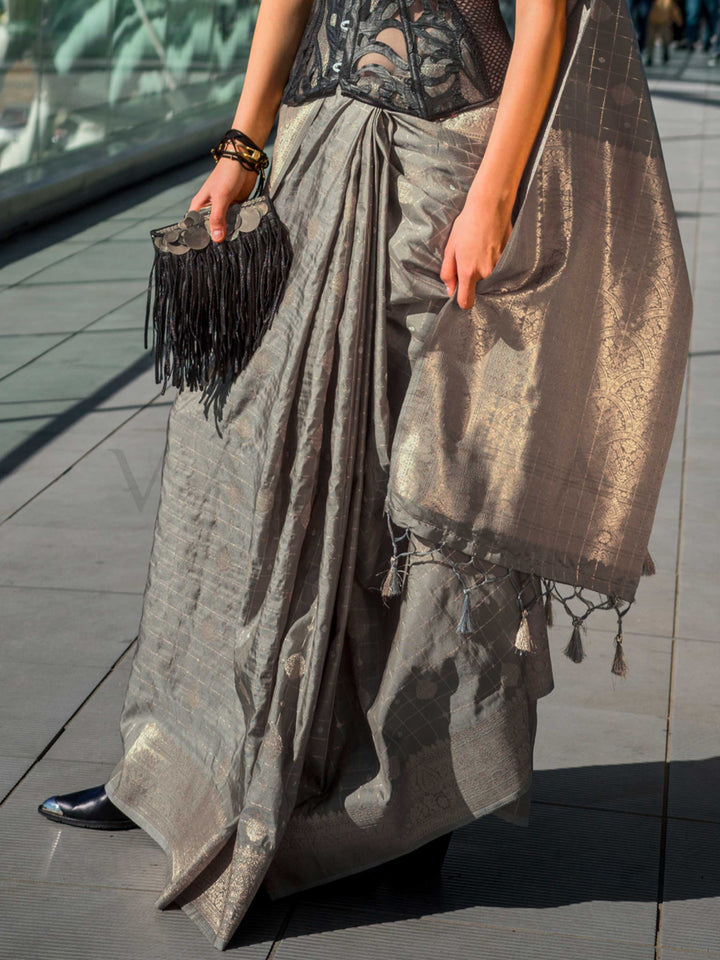 Champagne Grey Katan silk saree with Matte Silver zari detailing, featuring checks handloom weaving, designer blouse and tasselled pallu — a perfect fit for bollywood and award nights. Styled with modern belts, hats, and accessories to blend heritage and couture elegance. This image captures the back drape focus revealing pallu finesse and motif richness, ideal for visualizing festive elegance, designer appeal, and modern styling inspiration. Perfect for online shoppers seeking detailed previews before purc