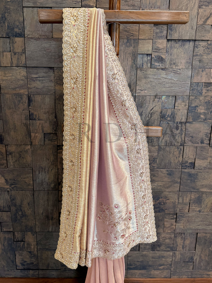Pink embroidered fabric draped over a wooden rod against a stone wall. Close-up of hand-embroidered border with floral zardozi motifs and red stones on cream tissue silk.