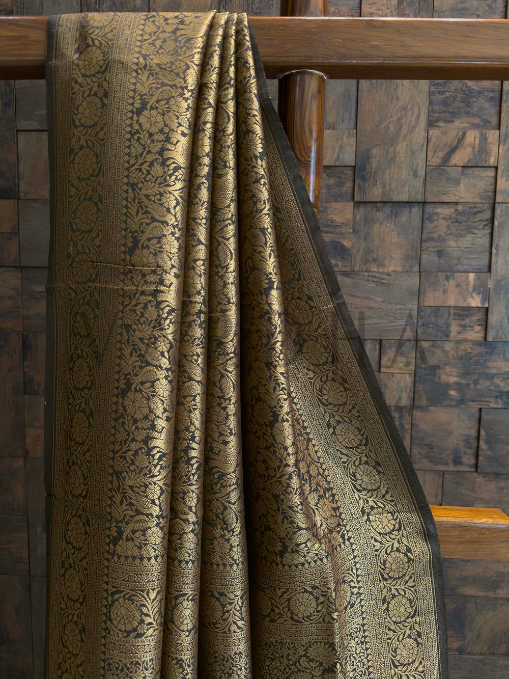 Textured fabric draped over a wooden chair against a wooden paneled wall.  Close-up of zari work on black Mysore silk saree featuring floral vines and creeper patterns