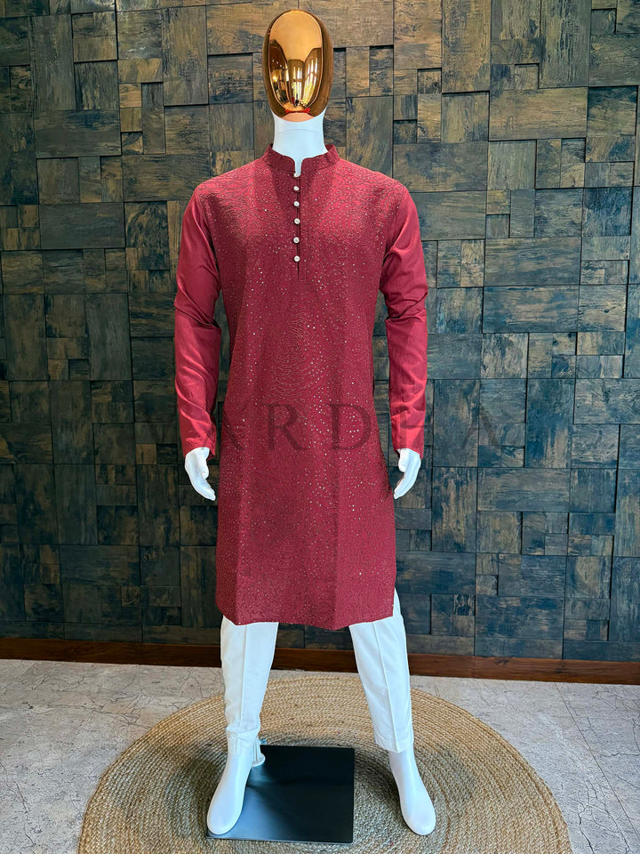Red kurta on a mannequin against a textured wall. Deep maroon viscose men’s kurta with mandarin collar and front panel covered in rosette swirl sequin embroidery, styled on mannequin with ivory churidar for festive wear.