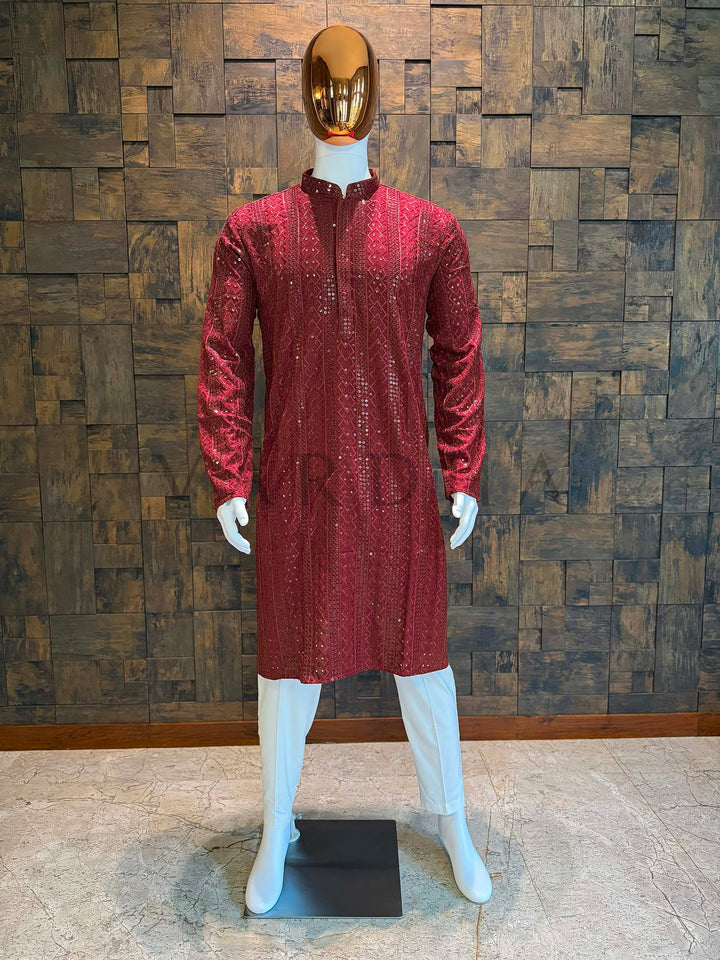 Mannequin wearing a red patterned outfit against a wooden paneled wall. Garnet red viscose men’s kurta with mandarin collar, concealed placket and vertical sequin-embroidered jacquard panels, styled on mannequin with white churidar for wedding wear.