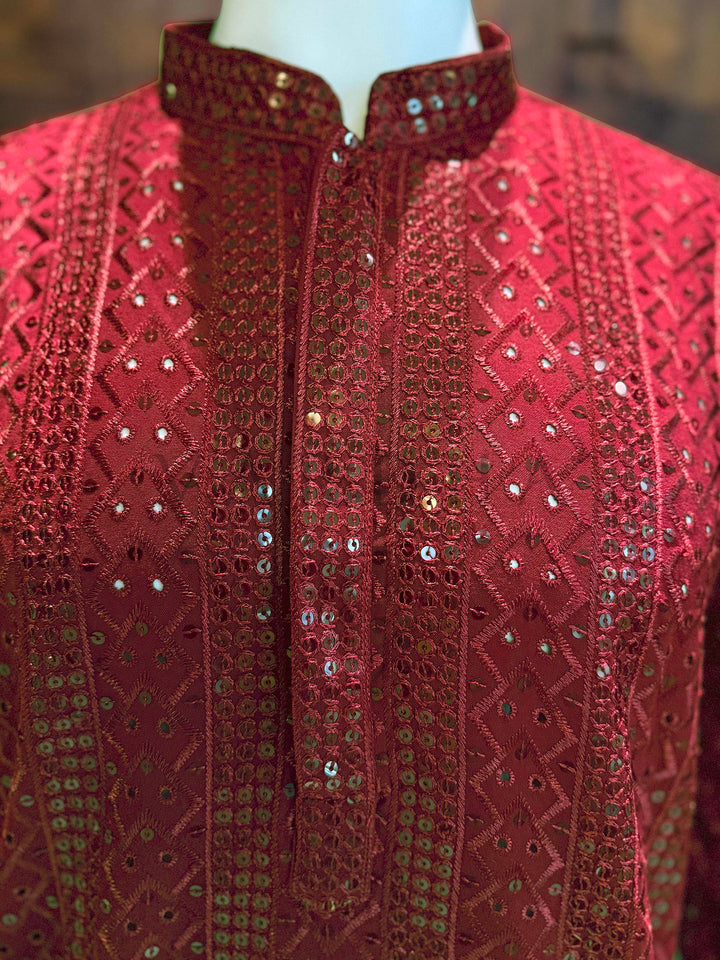 Red embroidered garment on a mannequin with a blurred background Close view of red kurta neckline showing sequin-edged band collar and clean placket with geometric jacquard texture and reflective sequin highlights.