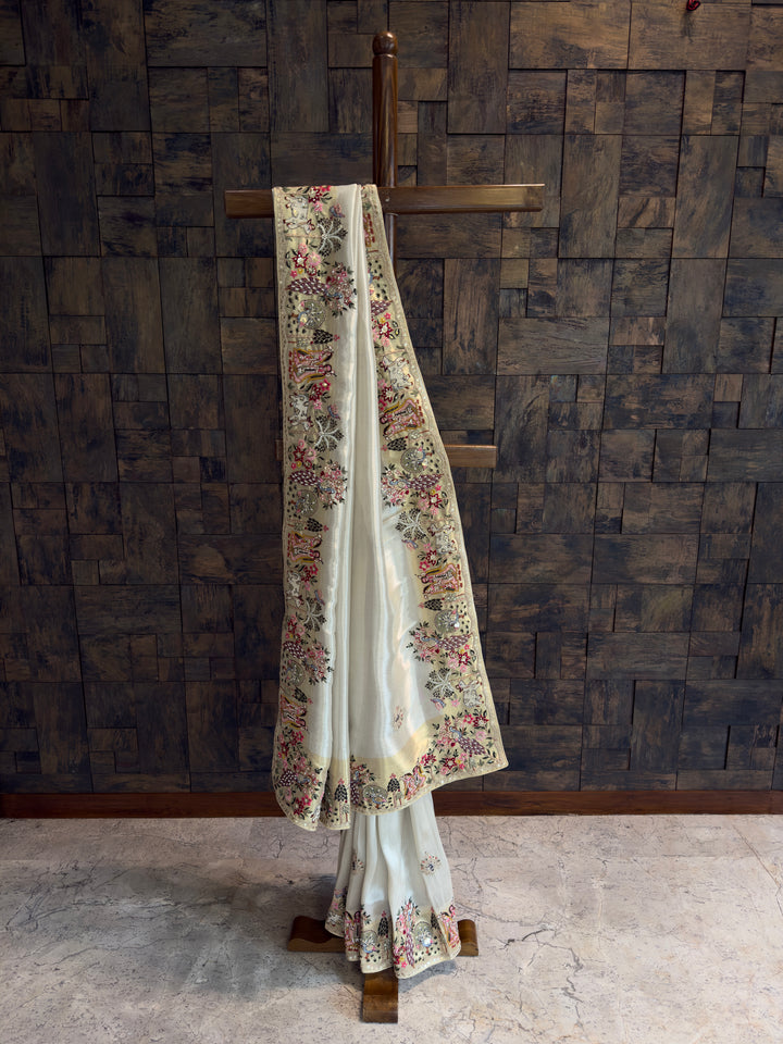 White saree with floral embroidery on a wooden stand against a textured dark wall.