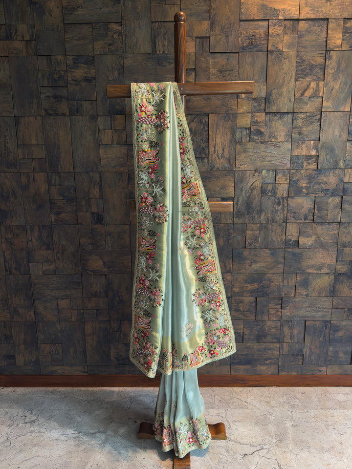 Pista green tissue art silk saree with gold undertone, hand embroidery, mirror work and zari, featuring Radha Krishna, peacocks, cows and floral trees on an ornate story border.