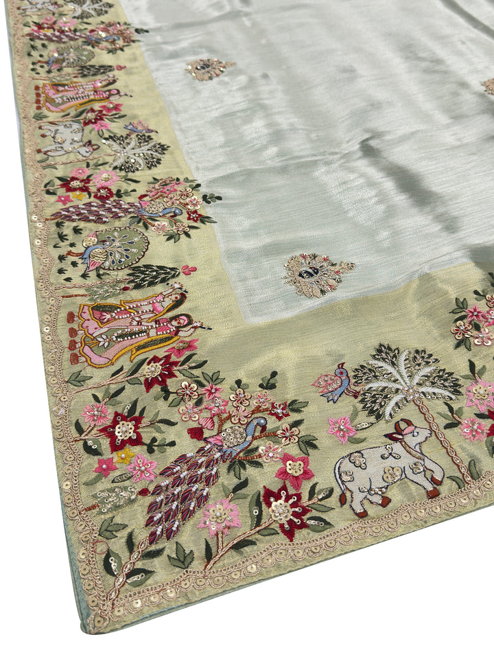 Dusty green tissue silk saree featuring Radha Krishna and peacock embroidery with floral zari motifs—ideal for traditional rituals and ceremonies.