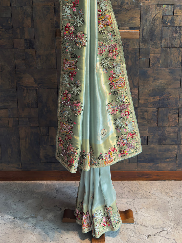 Sacred cow, birds and temple trees embroidered along the gold-tinted border of turquoise tissue saree; elegant storytelling design.