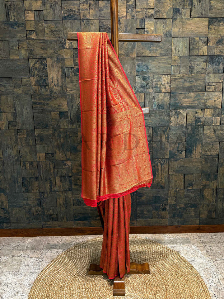 Orange saree on a wooden stand against a textured wall. Red Orange Mysore Silk Saree displayed on a wooden hanger with rich gold zari work glowing against a dual-tone base