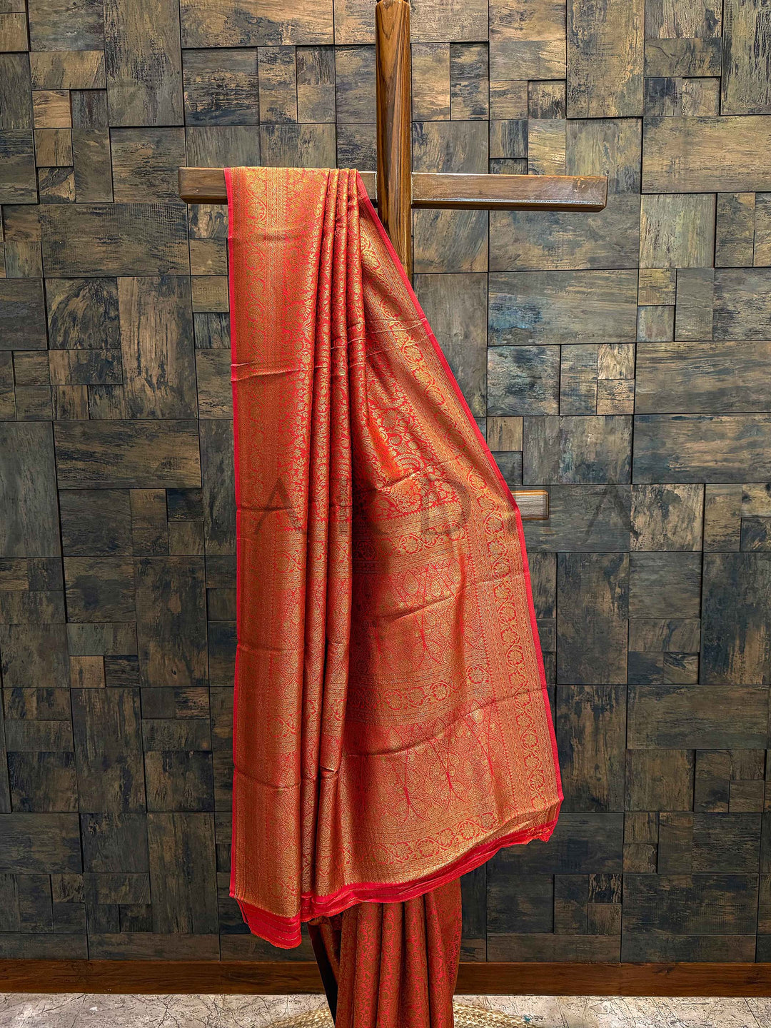 Orange saree with a red border draped over a wooden rod against a textured wall. Drape on natural jute base showcasing texture, color warmth, and fall of the Mysore silk
