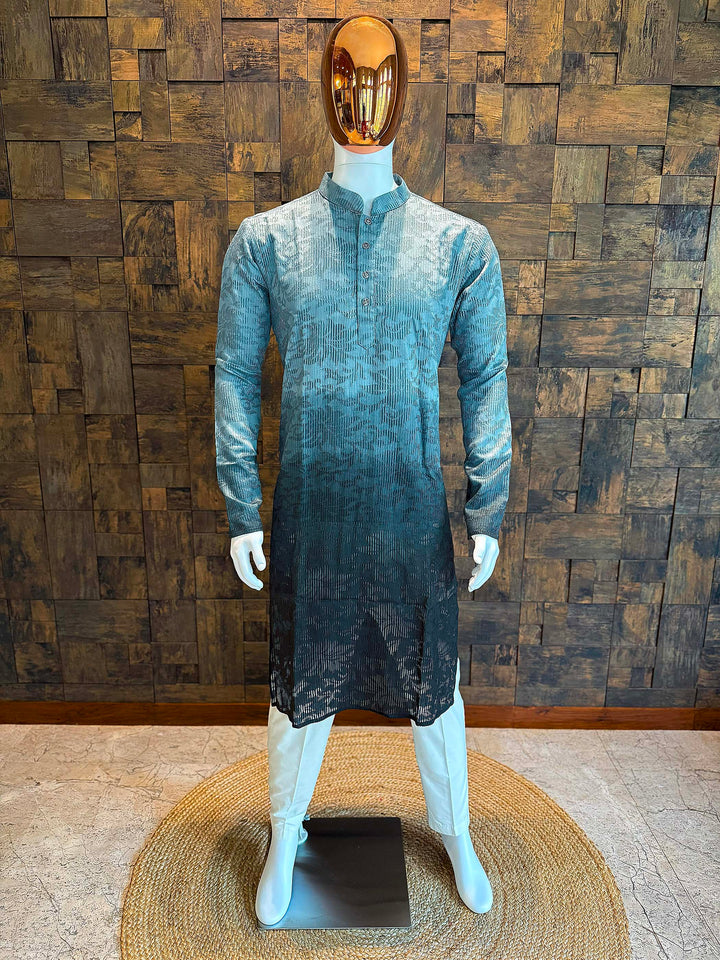 Mannequin wearing a long, textured dress against a wooden paneled wall. Teal Grey ombre viscose men’s kurta with mandarin collar and self-textured jacquard pattern, fading to deep charcoal at the hem, styled on mannequin with white churidar.