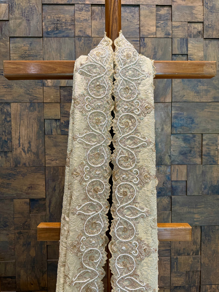Close-up of hand-done embroidery in paisley and floral motifs featuring golden zari and stone embellishments. Decorative lace scarf draped over a wooden cross against a wooden paneled wall.