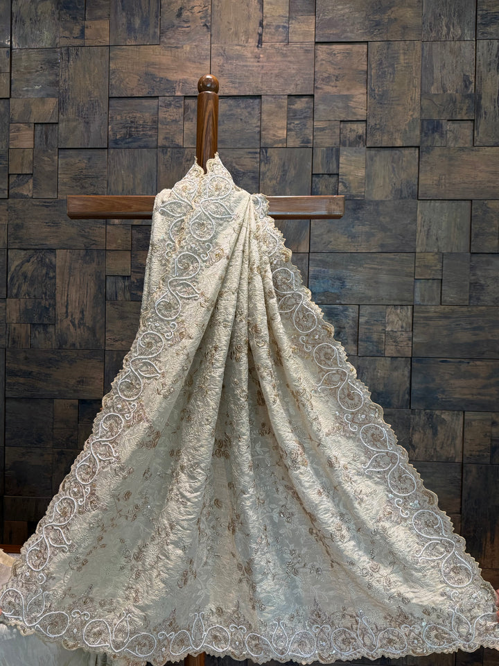 Saree pallu spread showcasing the ornate craftsmanship and light-reflective tissue silk texture. Lace fabric draped over a wooden cross against a wooden paneled wall.