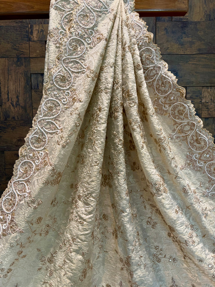 Detailed border with white beading, golden zardozi swirls, and delicate scalloped lace edge. Decorative fabric with intricate patterns draped over a wooden surface