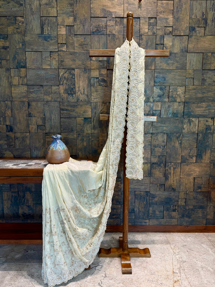 Full view of ivory saree with hand zardozi, sequins, and scallop stonework border on luxurious tissue base. Lace fabric draped over a wooden stand against a textured wall.