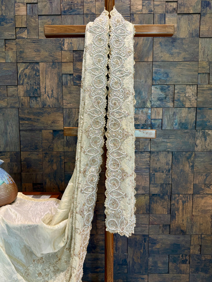 Ivory designer saree with heritage embroidery ideal for bridal events and special celebrations. Lace garment hanging on a wooden cross against a wooden paneled wall.