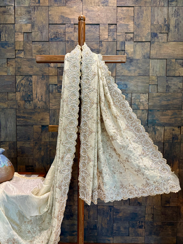 Tissue silk body in shimmer finish with floral motifs across the field of the saree. Luxurious bridal saree in ivory with full-length designer detailing perfect for receptions and sangeet.  Lace fabric draped over a wooden cross against a wooden paneled wall.