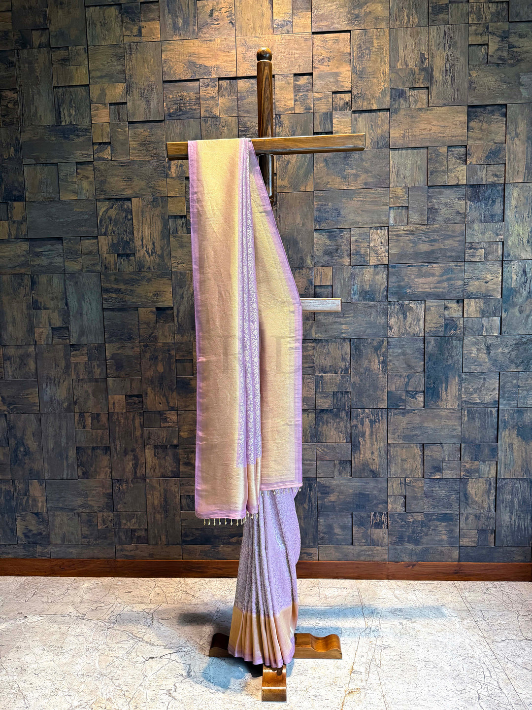 Saree draped over a wooden rod against a textured wall. Lilac and Silver  Kanjivaram Tissue Silk saree with floral jaal displayed on a wooden stand against a textured wall.