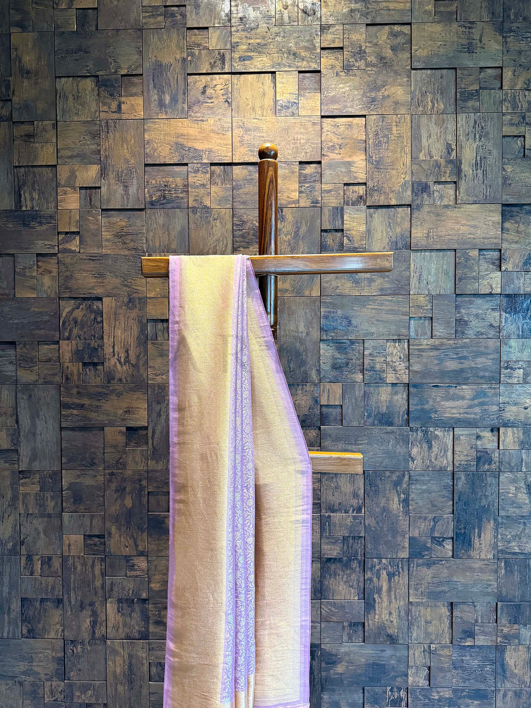 Wooden cross with a towel draped over it against a wooden paneled wall. Close-up of shimmering gold tissue border with lilac selvedge on Kanjivaram saree. Detailed view of lilac floral creeper jaal weaving on tissue silk fabric.