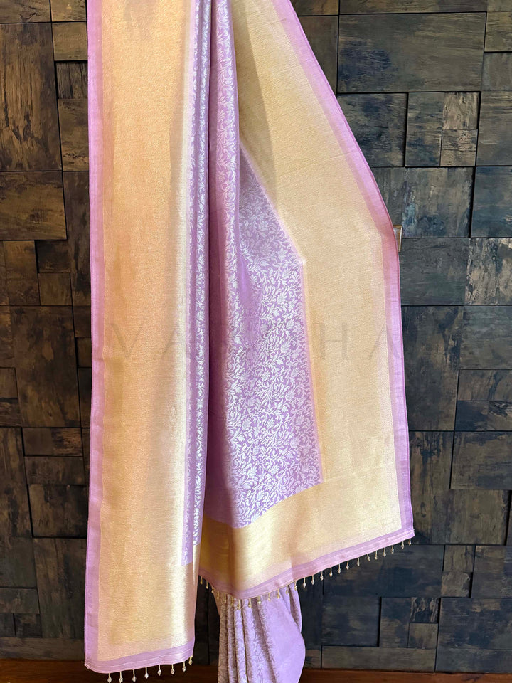 Light pink and beige fabric with lace detailing draped over a wooden surface. Pallu shot showing lilac woven panel framed with gold tissue and tassel finish. Full-length display of lilac–gold tissue silk saree highlighting soft sheen and border.