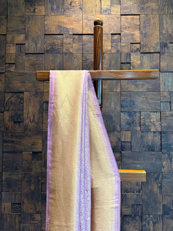 Wooden towel rack with a towel against a wooden paneled wall. Coordinated blouse piece with matching lilac tone and border placed with the saree. Edge view of saree showing tasselled finish and neat lilac selvedge.