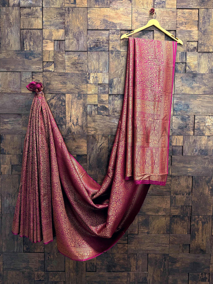 Pink saree with gold patterns draped over a wooden hanger against a wooden paneled wall. Wine maroon Mysore silk saree with golden zari floral brocade pattern draped elegantly against a wooden wall background