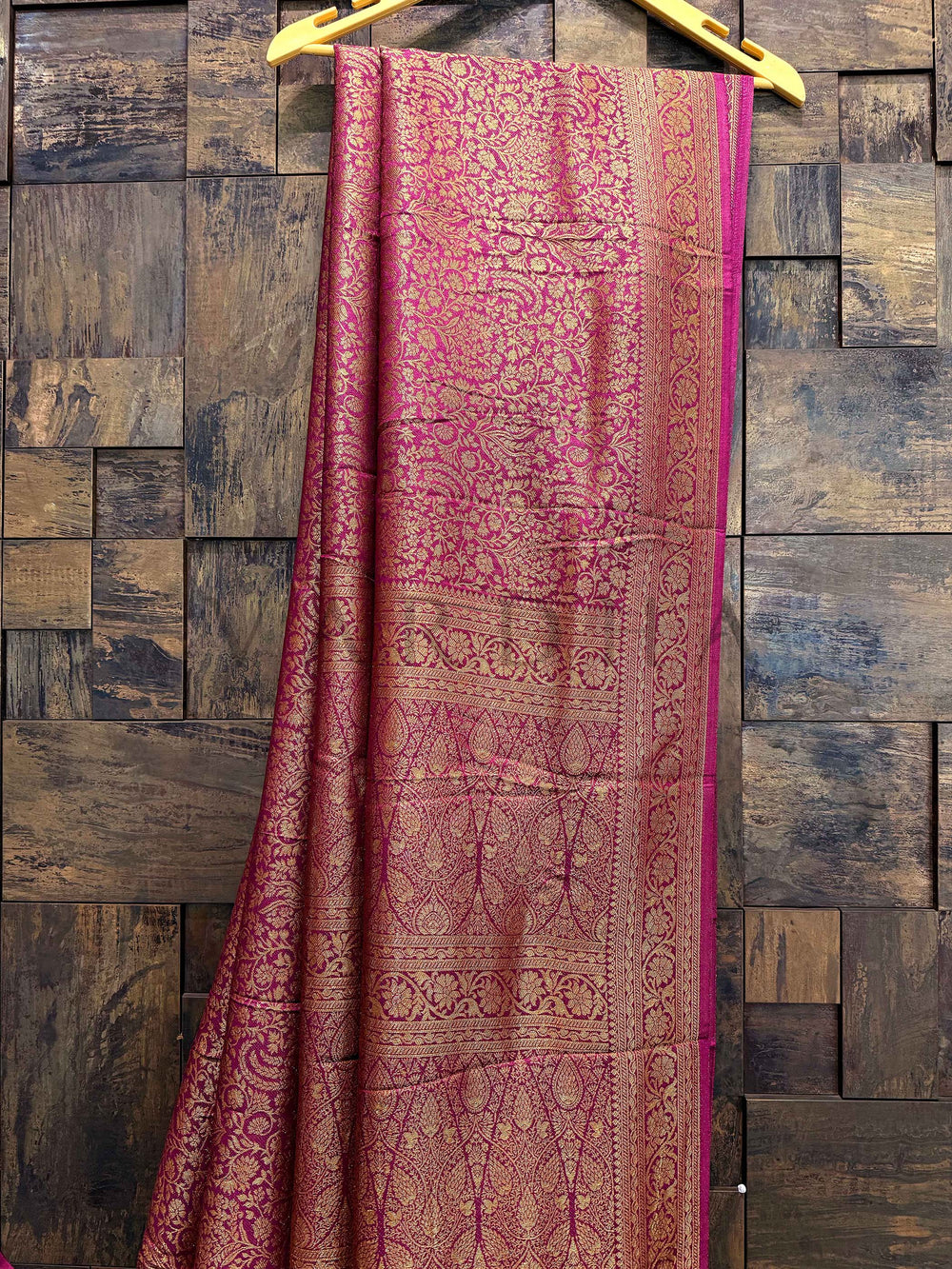 Pink and brown patterned fabric draped over a wooden hanger against a wooden paneled wall. Close-up of woven gold zari pallu with floral and temple motifs on wine Mysore silk fabric