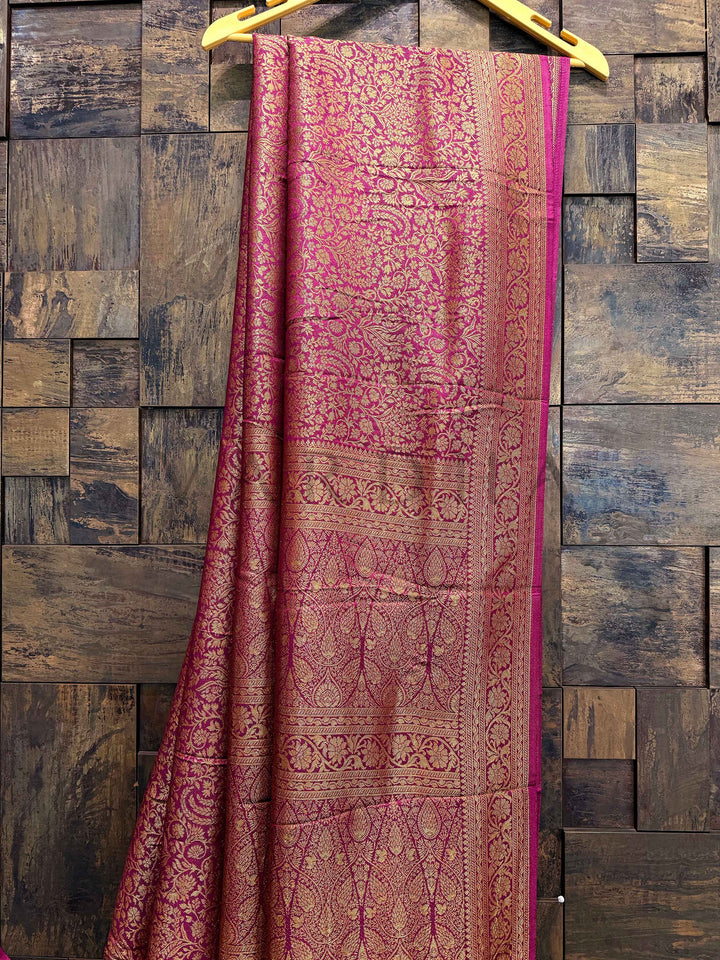Pink and brown patterned fabric draped over a wooden hanger against a wooden paneled wall. Close-up of woven gold zari pallu with floral and temple motifs on wine Mysore silk fabric