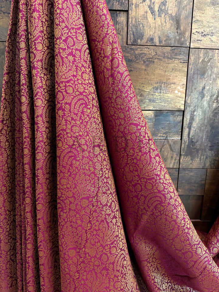 Pink and gold patterned fabric draped over a wooden surface Blouse piece attached in wine maroon shade with subtle zari hints complementing the saree body Draped pleats highlighting the shimmer and crisp fall of the wine Mysore silk saree with zari detailing