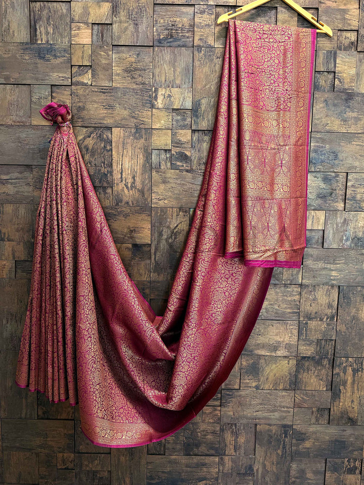 Pink saree with gold patterns draped over a wooden hanger against a wooden paneled wall. Zari border view of the saree showing symmetrical alignment and finishing across wine maroon base