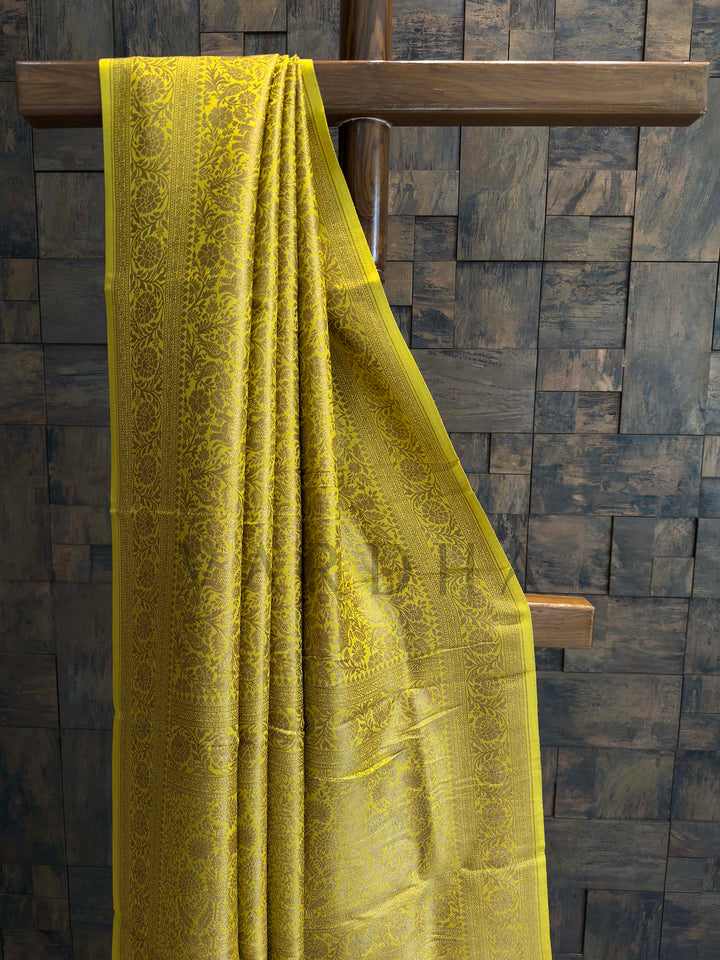 Yellow fabric draped over a wooden rod against a textured wall. Close-up of zari detailing on mustard yellow Mysore silk saree showing intricate floral creepers and motifs.