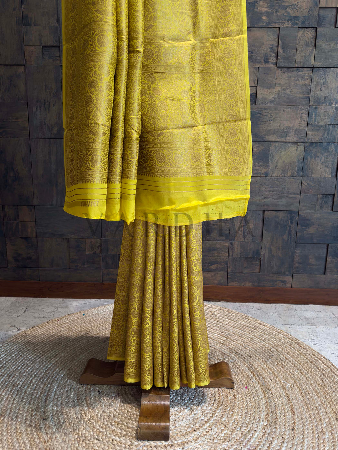 Yellow saree with a textured pattern on a wooden stand against a stone wall. Pleats of mustard yellow Mysore silk saree enriched with golden zari elephant and parrot motifs.