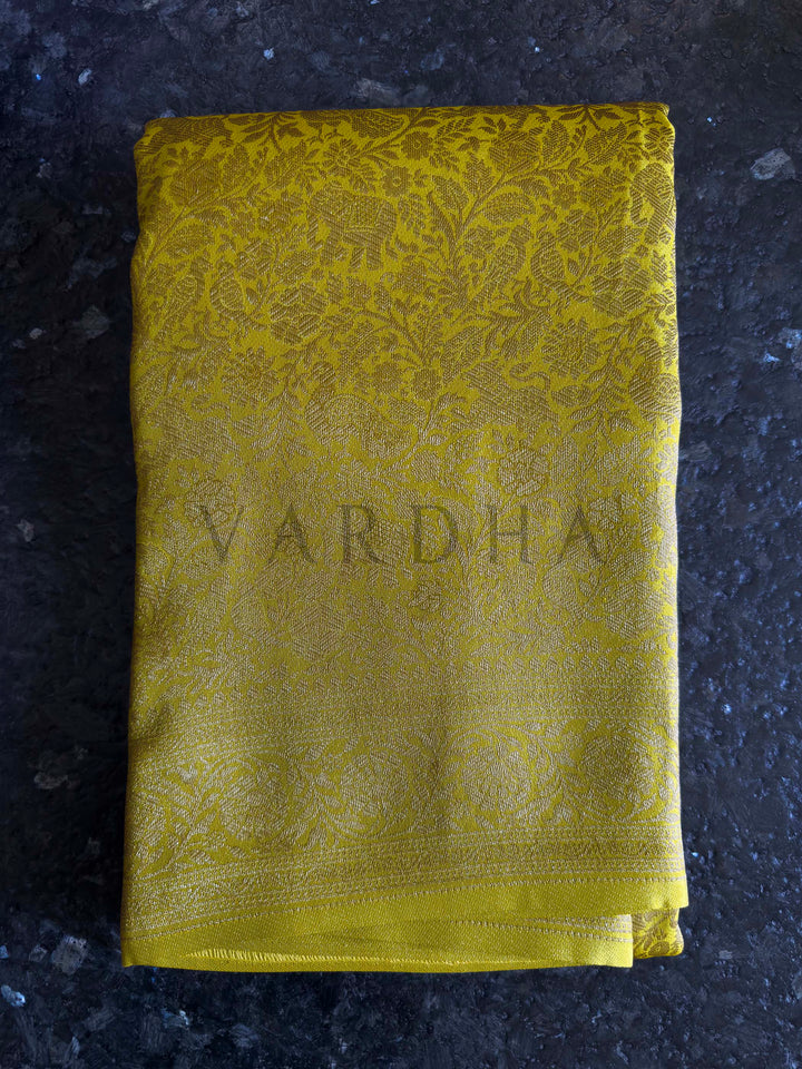 Yellow fabric with 'Vardha' branding on a dark textured surface Elephant and peacock motifs woven in gold zari on mustard yellow Mysore silk fabric.