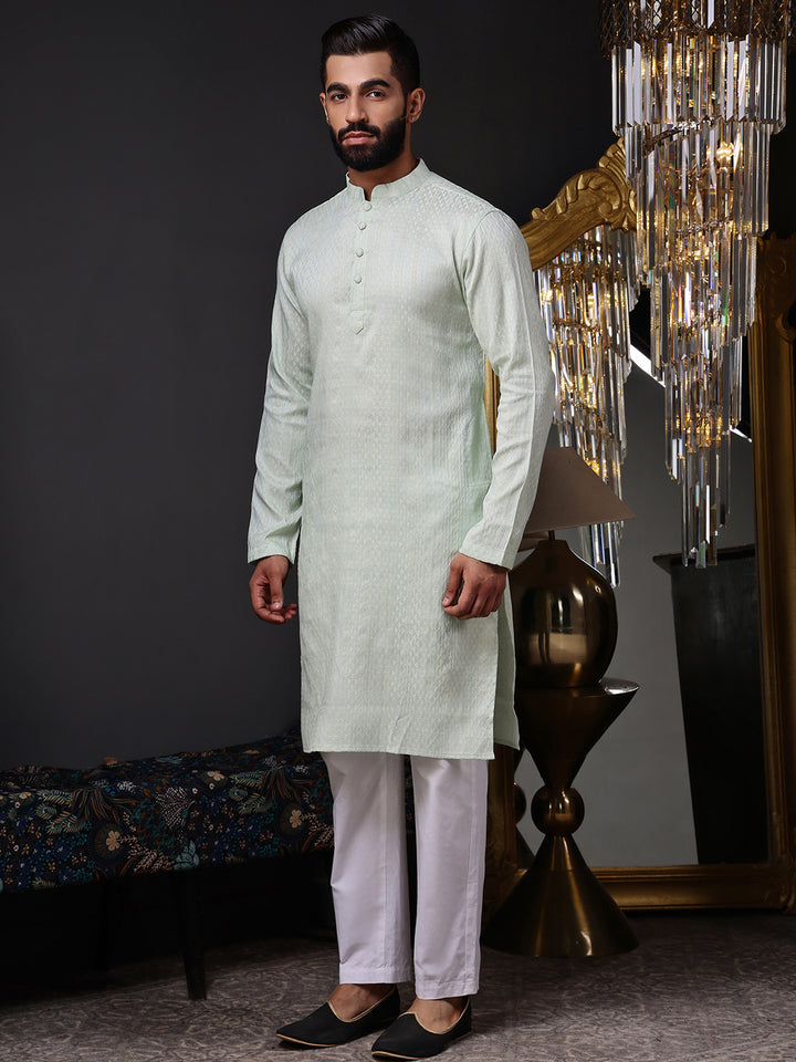 Man wearing a light green kurta with white pants in an elegant room setting. Shop our modern mint green kurta with a sophisticated self-textured design for a fresh ethnic look