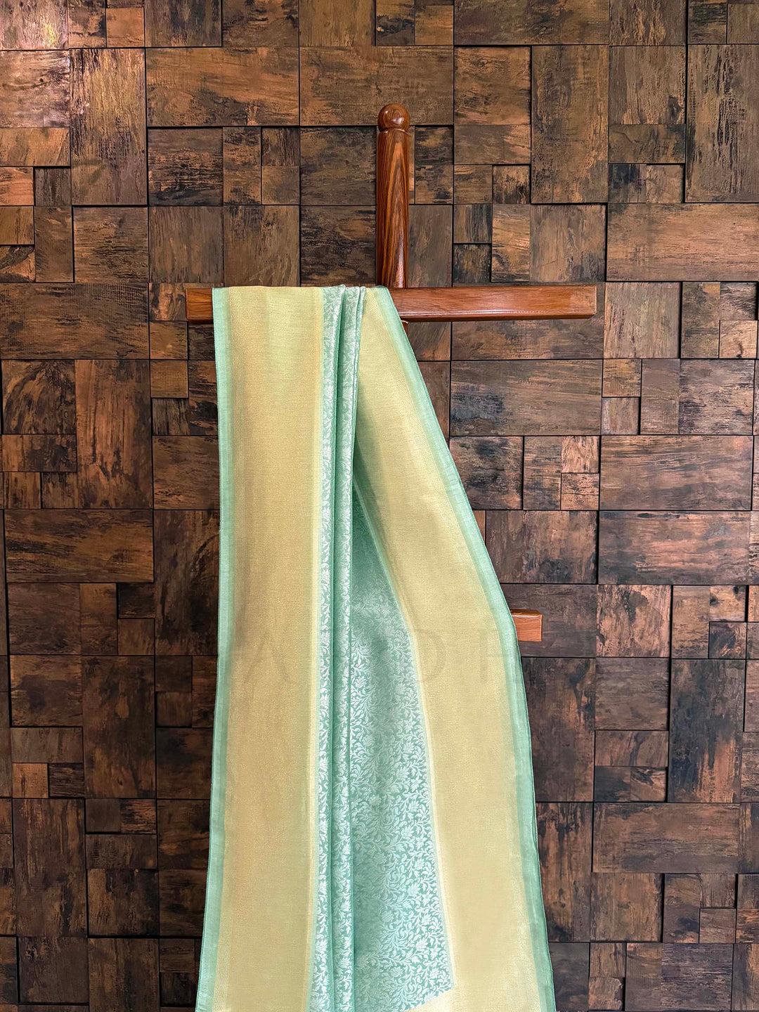 Green and beige towel draped over a wooden hanger against a wooden paneled wall. Seafoam green and butter-gold Kanjivaram Tissue Silk saree with floral jaal displayed on a wooden stand.