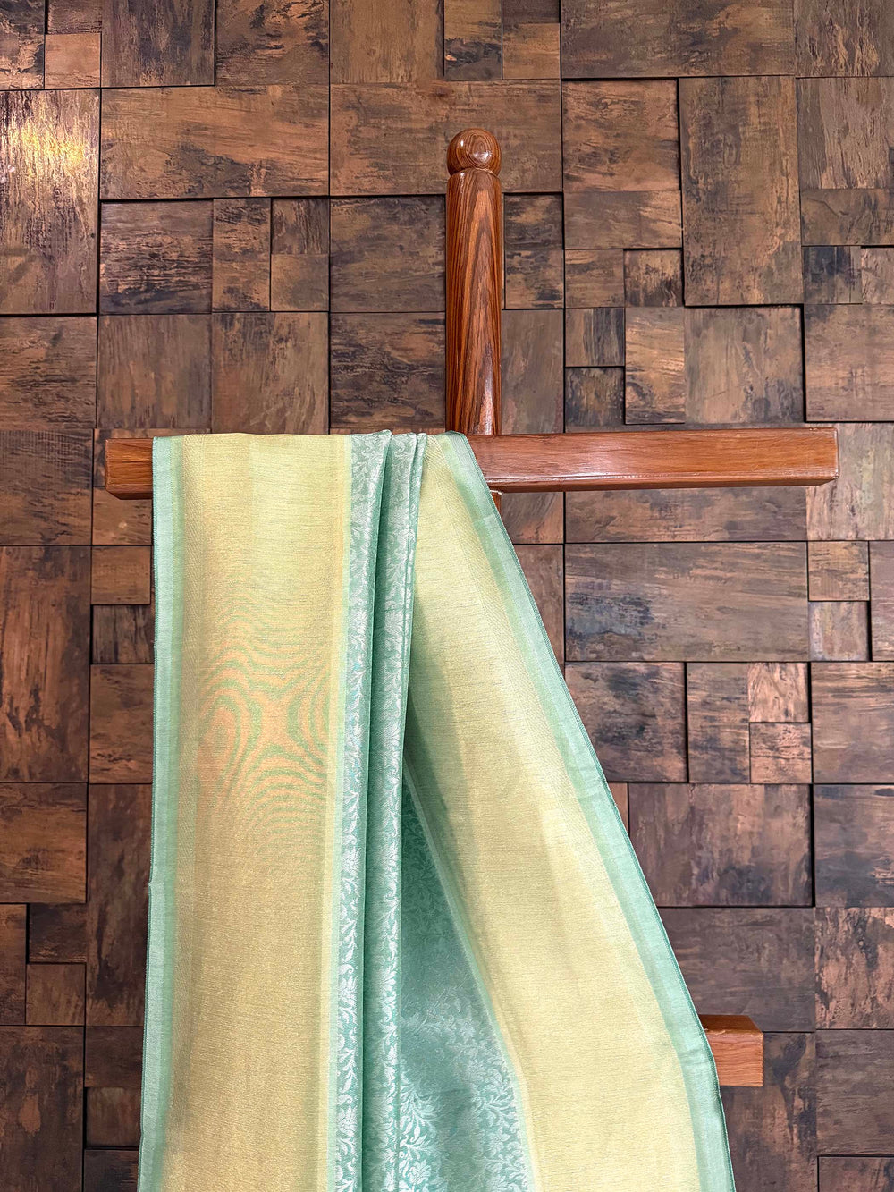 Wooden hanger with a green towel against a wooden paneled wall Close-up of shimmering butter-gold tissue border with seafoam selvedge on Kanjivaram saree.