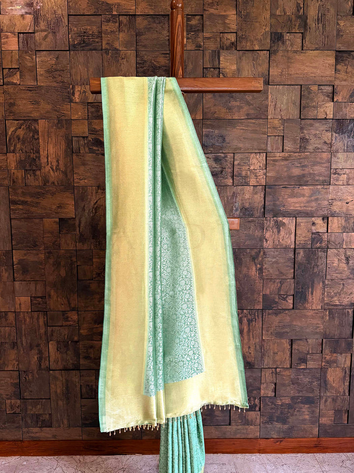 Green and beige saree draped over a wooden hanger against a wooden paneled wall. Pleated section of saree emphasizing fluid fall and delicate jaal weave.