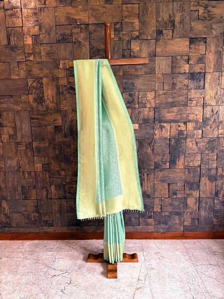 Light green saree on a wooden stand against a wooden paneled wall. Coordinating blouse piece shown with saree, matching sheen and border detail. Full-length view highlighting two-tone glow of seafoam body and gold border.