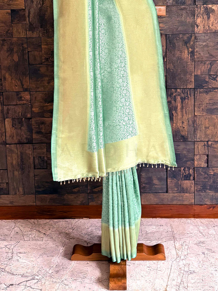 Green and beige fabric draped over a wooden stand against a wooden paneled wall. Edge view capturing neat tassels and refined zari tissue frame. Pallu shot showing luminous tissue finish with tasselled edging.