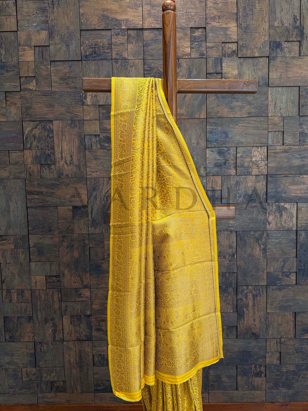 Yellow saree draped over a wooden hanger against a textured dark wall. Traditional border detailing in close-up showing zari vines and scroll patterns Pleats captured to show texture fall and refined golden motif arrangement