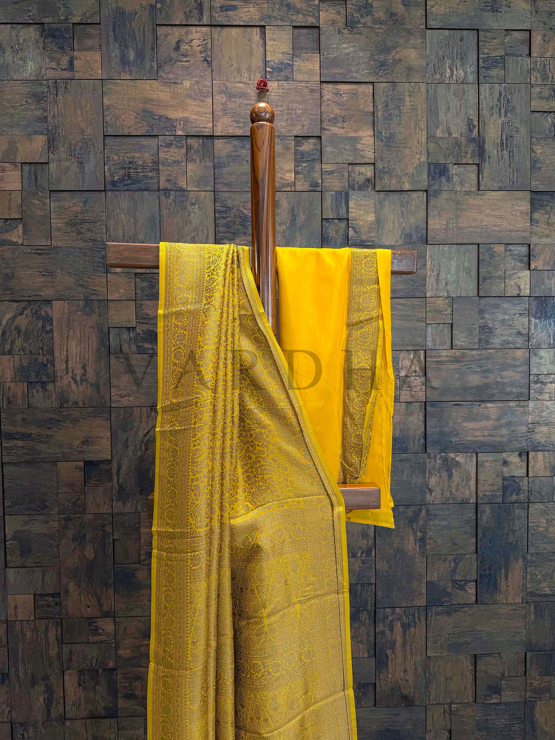 Yellow fabric with a brand logo draped over a wooden rod against a textured wall. Close-up of zari work on saree pallu with detailed floral and geometric patterns