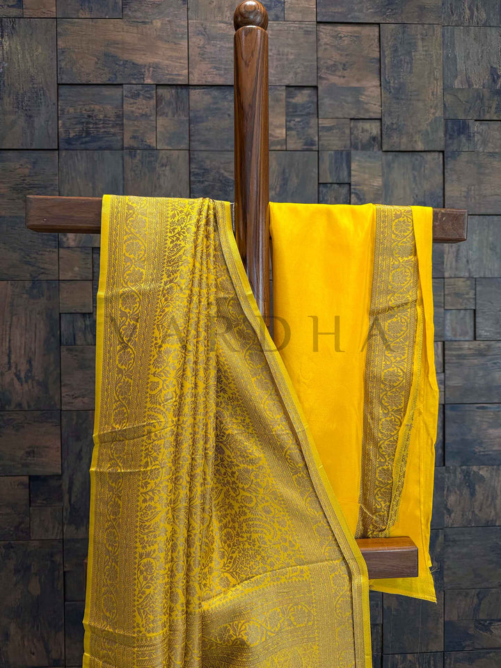 Contrast blouse fabric in matching mustard with zari border displayed behind saree. Yellow fabric with intricate patterns hanging on a wooden rod against a textured wall. Saree pallu drape showcasing intricate Mysore style zari craftsmanship