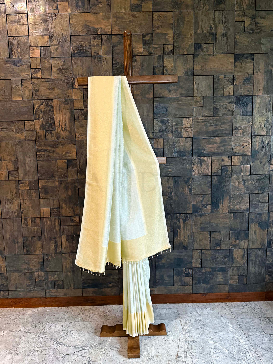 Beige saree draped over a wooden stand against a textured brown wall. Pistachio-mint or off white and butter-gold Kanjivaram Tissue Silk saree displayed on a wooden stand against a textured wall.