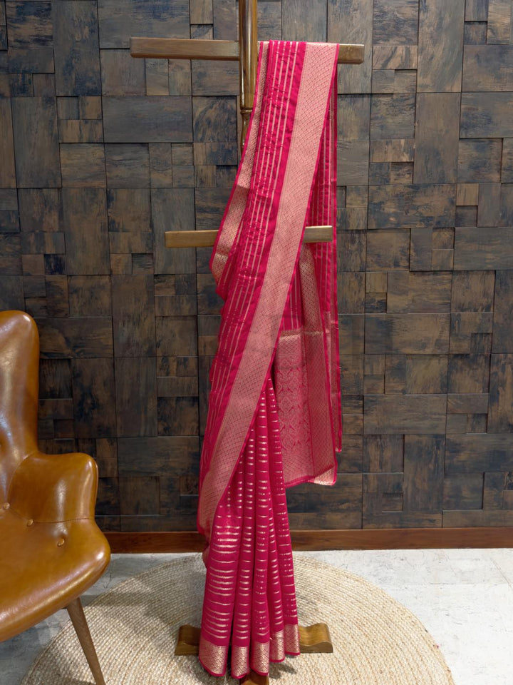 Rani pink feathersoft georgette saree with striped golden zari accents, styled on a wooden hanger for festive elegance., Close-up of zari striped pallu in gold over rich rani pink base with textured weave, ideal for haldi or reception events