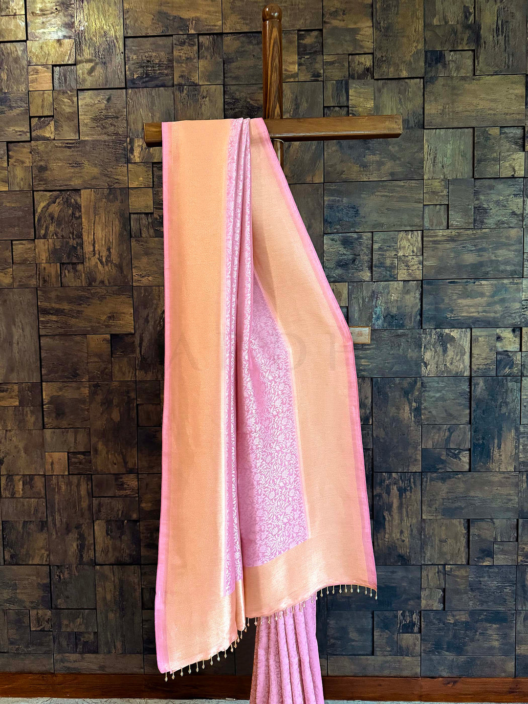 Pink saree with a wooden hanger against a textured wall. Rose pink and peach-gold Kanjivaram Tissue Silk saree with floral jaal displayed on a wooden stand.