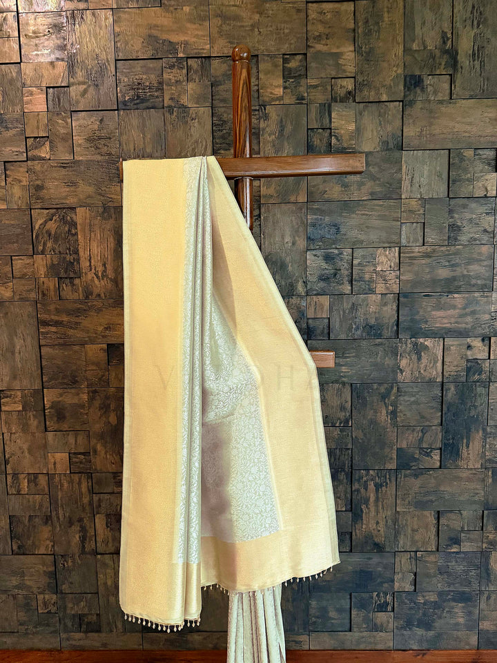 Beige and white saree draped over a wooden hanger against a textured brick wall. Full-length display of icy-mint Kanjivaram Tissue Silk saree showing two-tone glow. Edge view capturing refined zari tissue border and tasselled finish. Pleated section emphasizing fluid fall and delicate jaal weave.