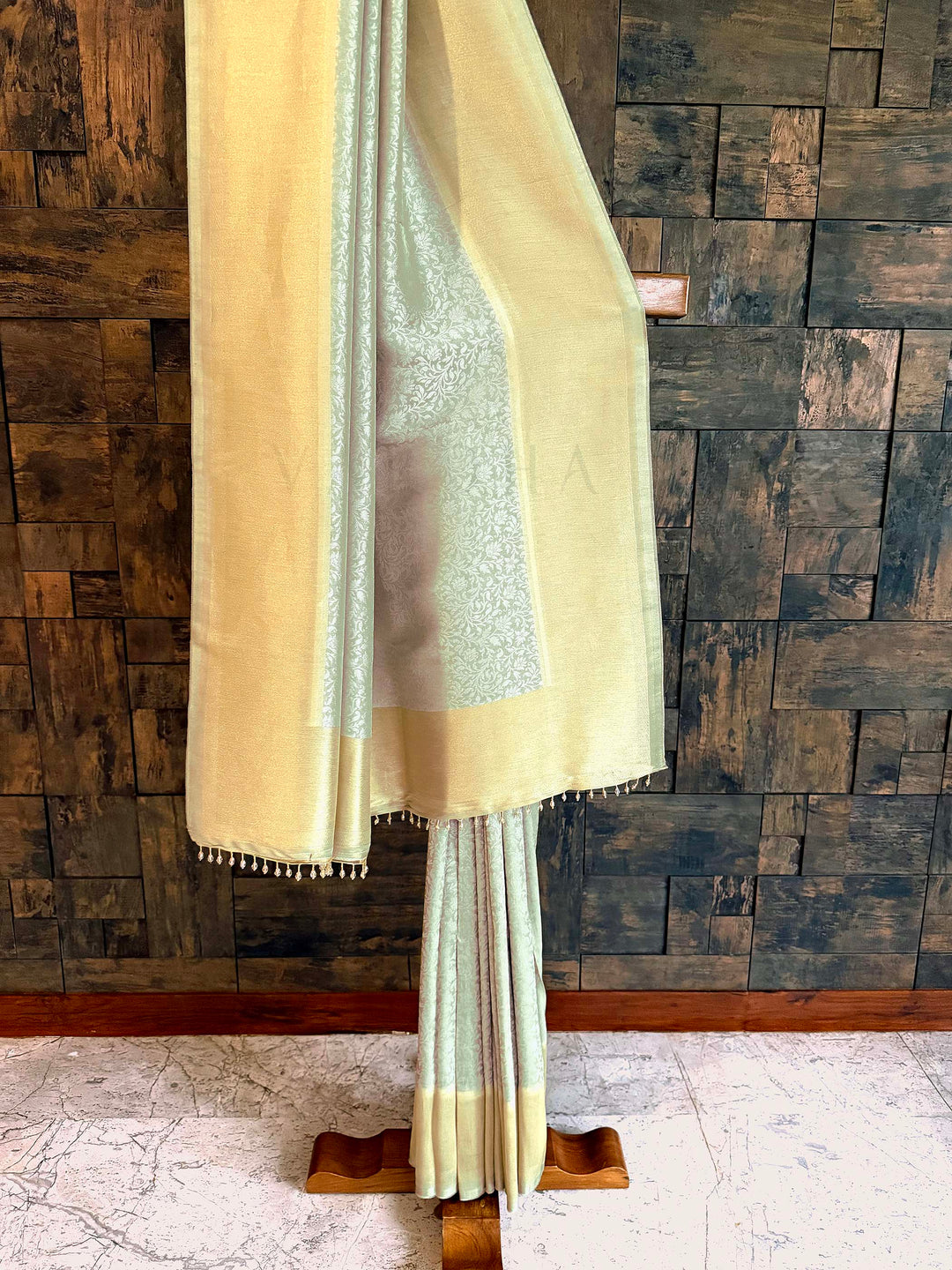 Beige saree with a textured pattern draped over a wooden stand against a wooden paneled wall. Pleated section emphasizing fluid fall and delicate jaal weave. Close-up of shimmering tissue border in butter-gold with icy-mint selvedge. Pallu shot highlighting luminous tissue finish and neat tassel edging.