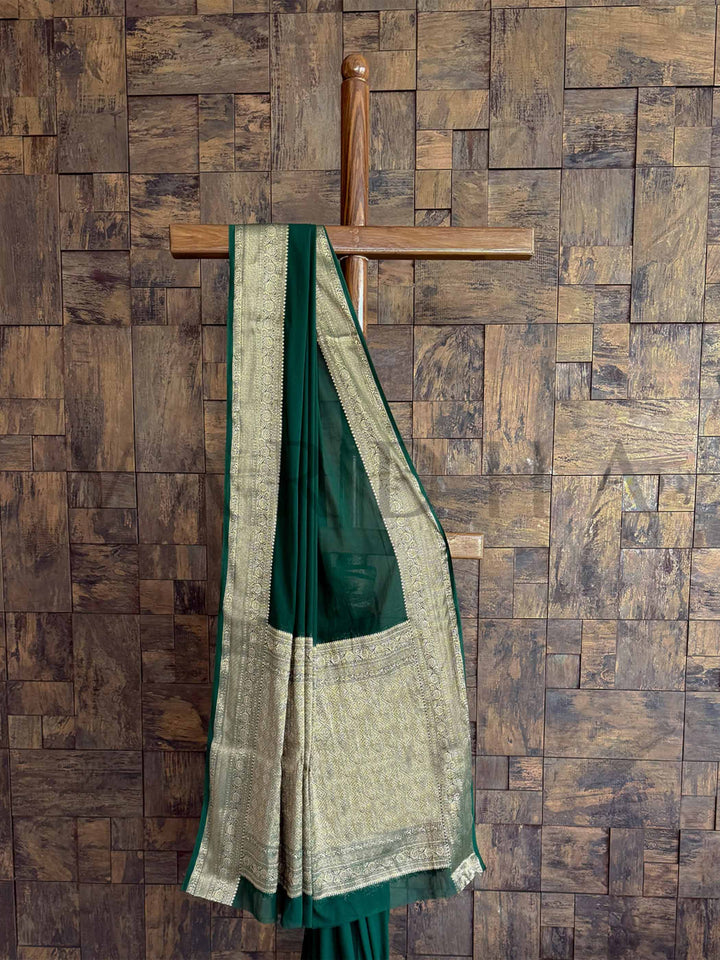 A luxurious deep green georgette saree with intricate gold zari embroidery, beautifully displayed to highlight its elegance. The soft, lightweight fabric enhances its graceful drape, ideal for celebrations.