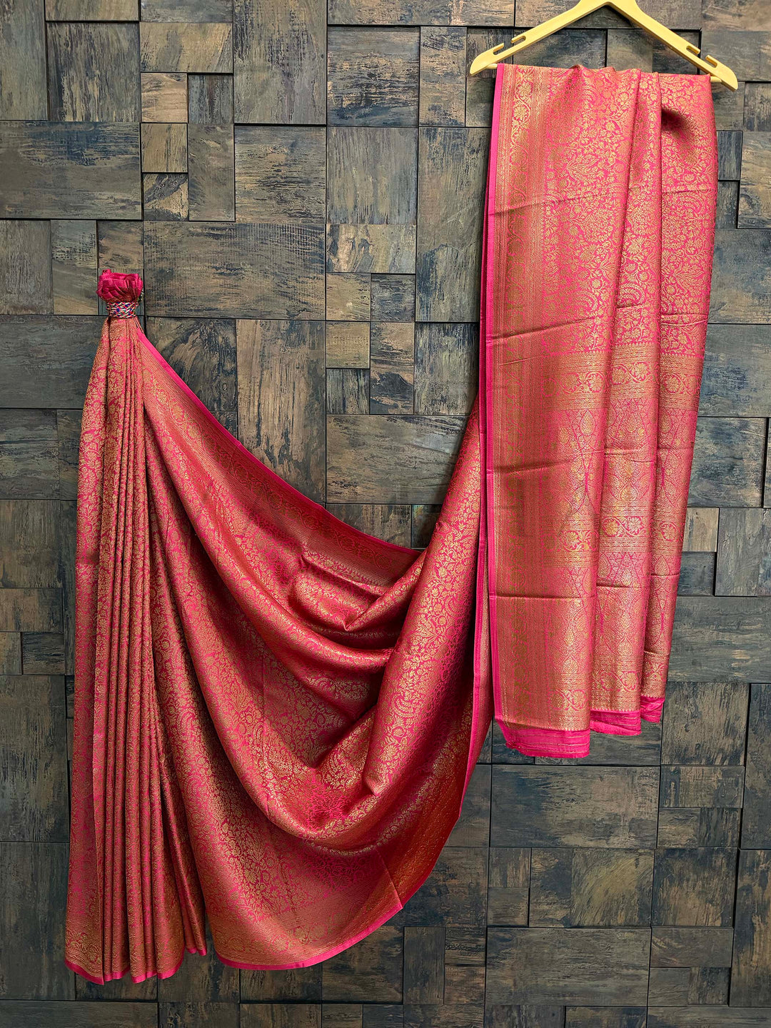Pink saree with gold patterns draped over a wooden wall. Rani Pink Mysore Silk Saree with golden zari brocade, gracefully draped for a festive look
