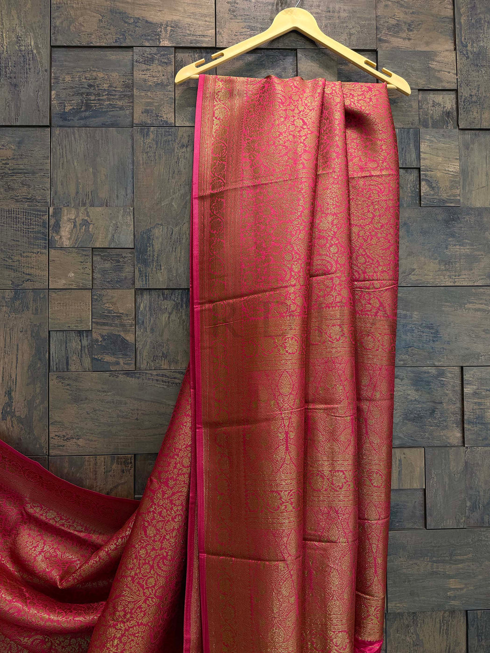 Red saree with gold patterns on a wooden hanger against a wooden.paneled wall. Close-up view of golden floral zari weave on soft viscose silk saree in pink
