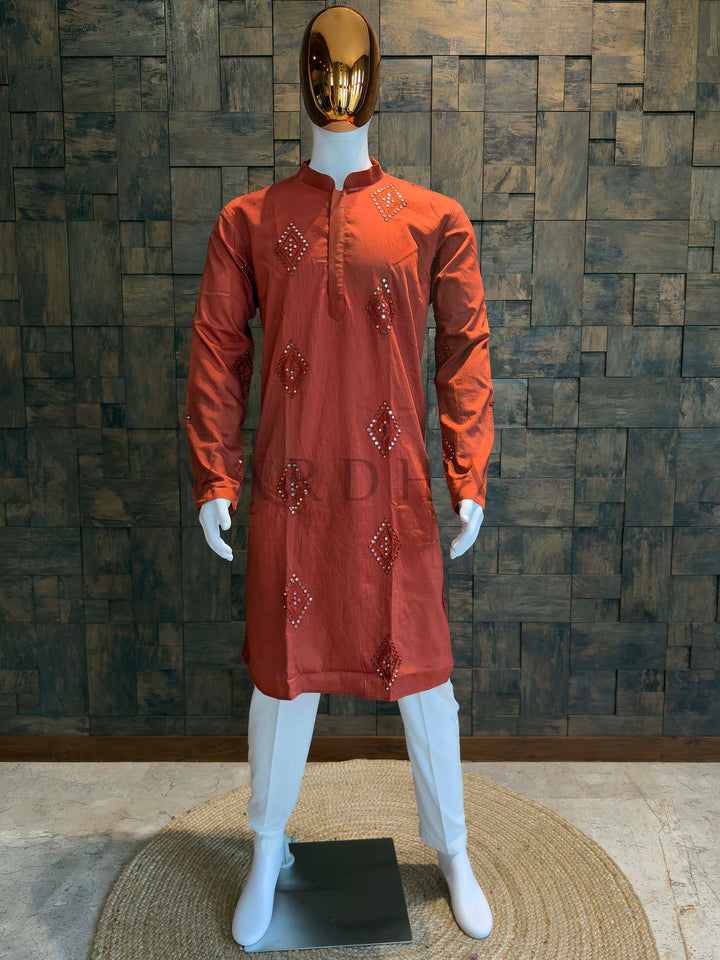 Orange kurta with embroidery on a mannequin against a wooden paneled wall. Rust orange viscose men’s kurta with mandarin collar, concealed placket and diamond mirror (sheesha) butis, displayed on mannequin for festive occasions.