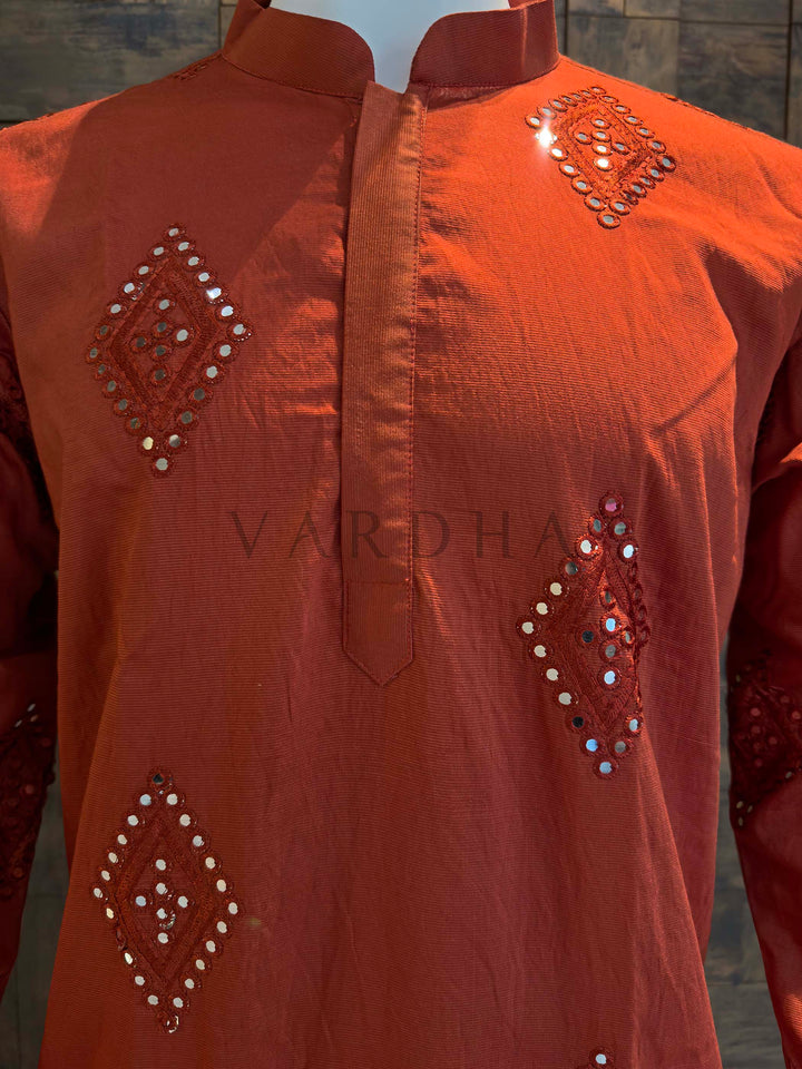 Orange embroidered shirt with mirror work on a plain background Close-up of rust kurta neckline showing band collar and neat placket with shimmering mirror embroidery arranged in geometric diamonds.